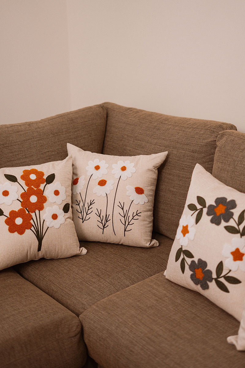 Beige cushion with embroidered white daisies and orange centers on a blue sofa – Autumn Bloom Collection