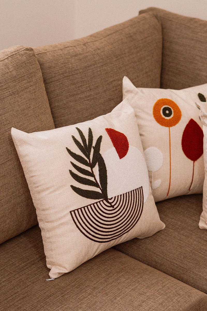 Beige cushion with embroidered palm leaf and geometric shapes in rust and green on a blue sofa – Autumn Bloom Collection