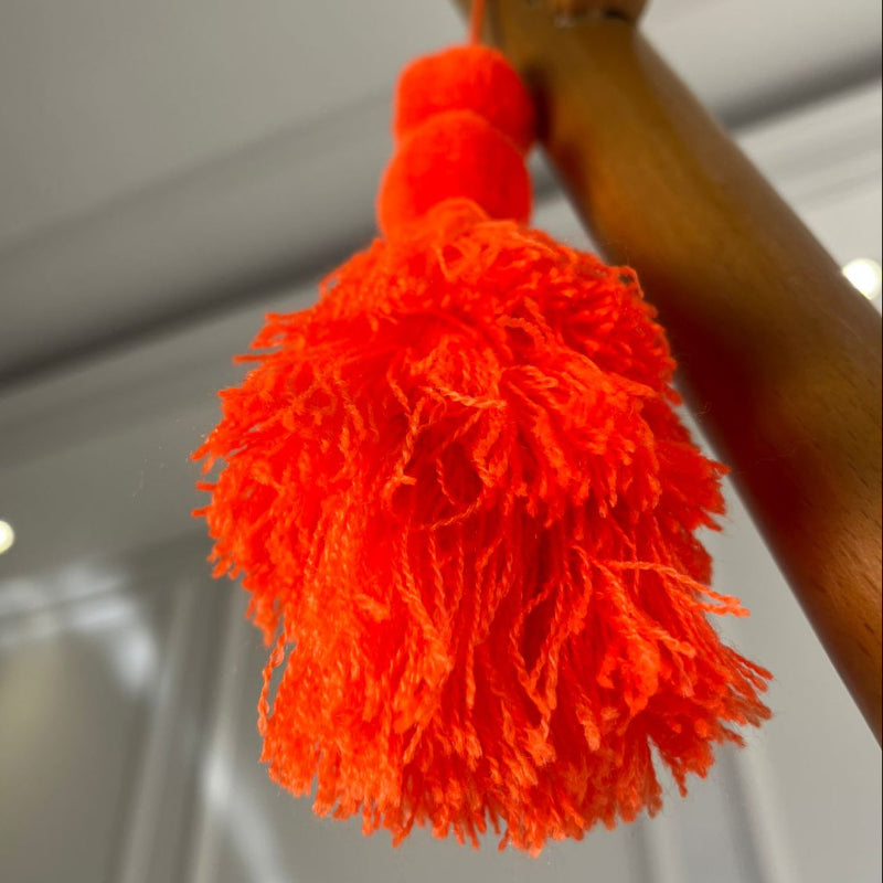 Neon orange pom-pom tassel hanging decoration with two pom-poms and layered neon orange yarn tassels hanging on a wooden chair.