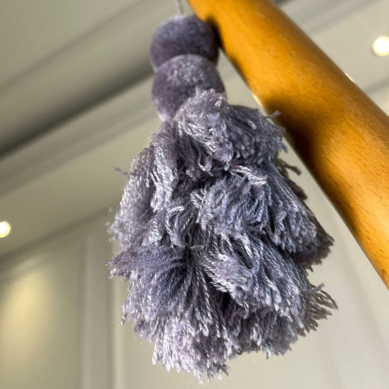 Grey pom-pom tassel hanging decoration with two pom-poms and layered grey yarn tassels hanging on a wooden chair.