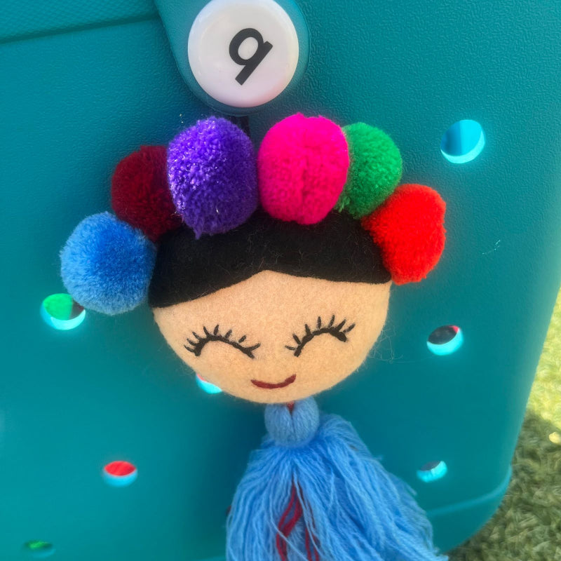 Handmade smiling Frida-inspired doll charm with colorful pom-poms and blue tassel on teal bag
