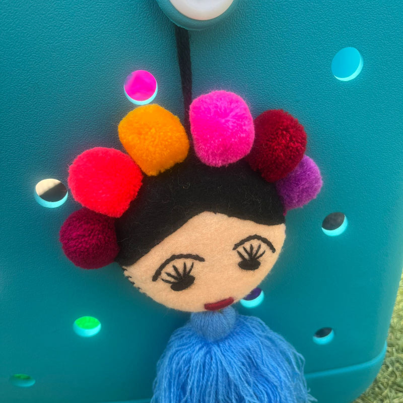 Handmade Frida-inspired doll ornament with colorful pom-poms and blue tassel attached to teal bag