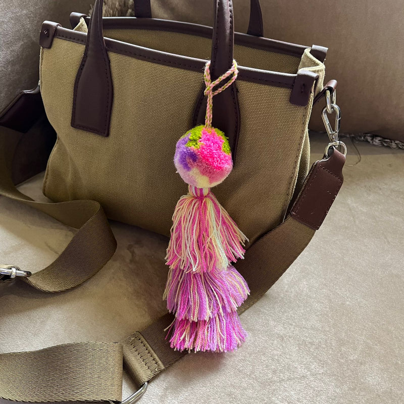 Soft pink pom pom tassel bag charm with layered pastel tassels attached to a beige handbag