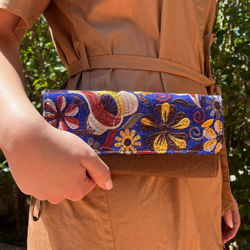 Hand holding a handmade brown leather clutch with colorful floral embroidery and wrist strap outdoors