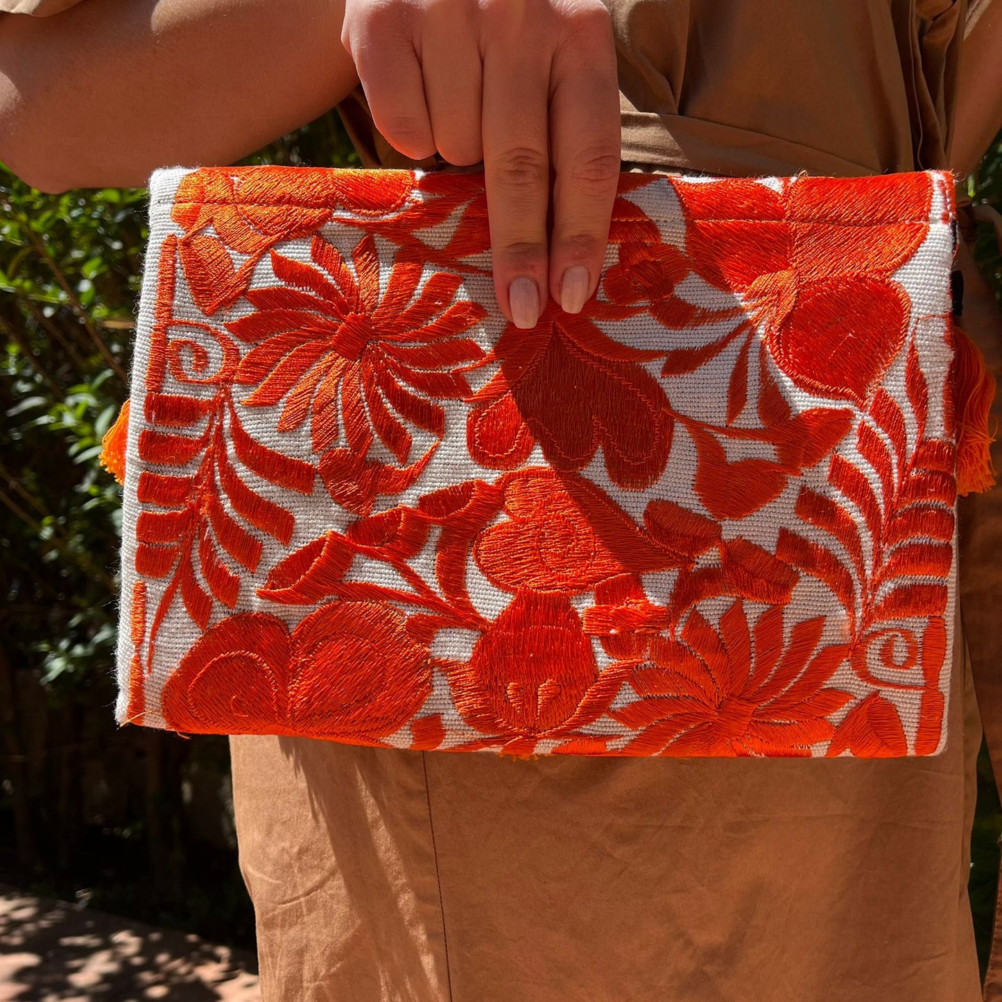Bright orange floral embroidered clutch bag with tassels held by a person wearing a brown dress outdoors