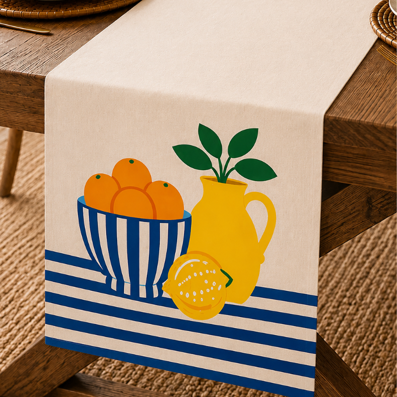 A cream table runner with a blue and yellow fruit and pitcher print, styled on a wooden dining table with plates, a glass, and a floral vase.