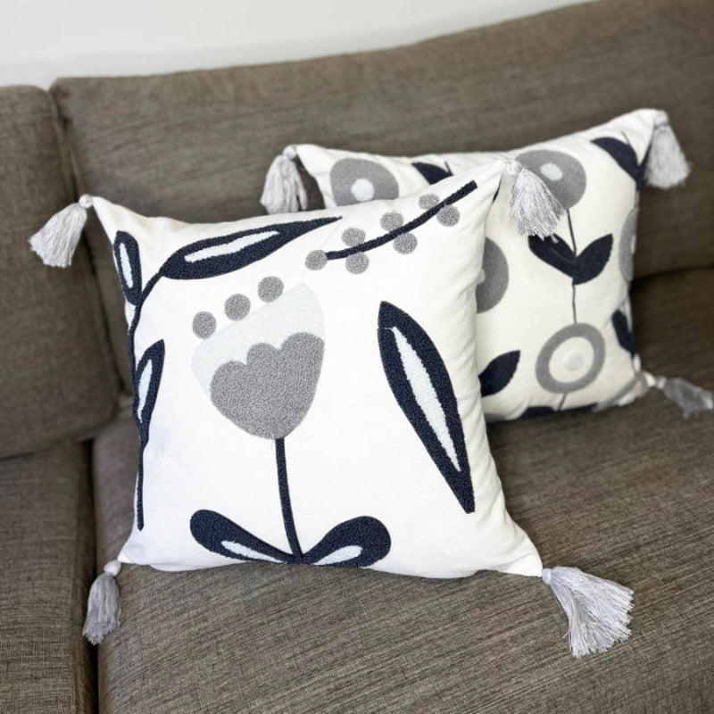 Embroidered floral cushion with navy and grey design, soft cream fabric, and tassel accents on each corner, perfect for home decor."