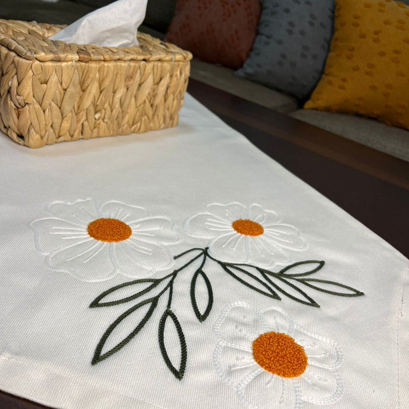 White cotton table runner with embroidered daisy flowers and orange centers on wooden coffee table