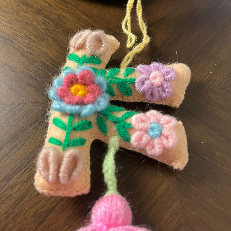 Handmade embroidered letter F ornament with floral patterns on beige fabric and pink tassel on wooden background