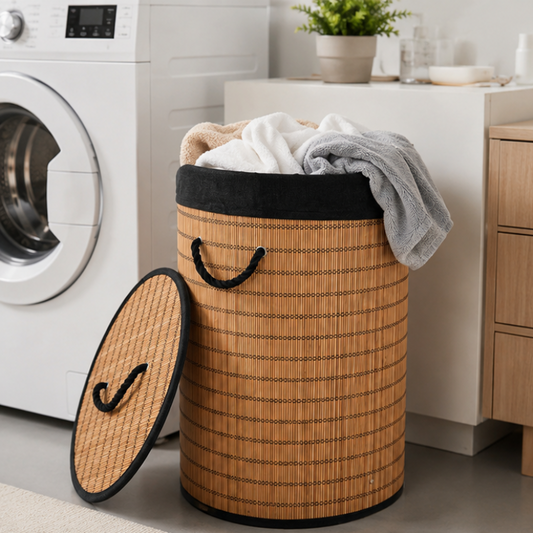 A bamboo-style laundry hamper with black trim and handles sits on a floor beside a washing machine, with folded white and gray towels draped over the top. The matching lid rests beside the hamper.