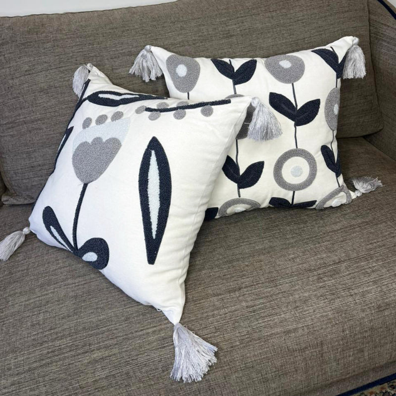 Embroidered floral cushion with navy and grey design, soft cream fabric, and tassel accents on each corner, perfect for home decor."