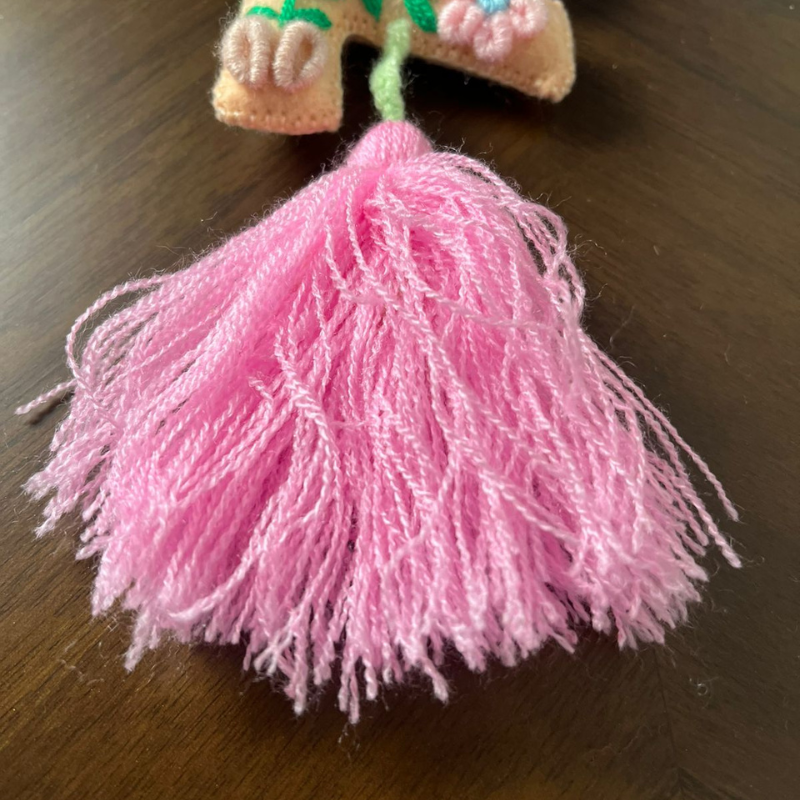 Handmade embroidered letter F ornament with floral patterns on beige fabric and pink tassel on wooden background