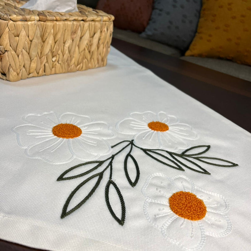 White cotton table runner with embroidered daisy flowers and orange centers on wooden coffee table