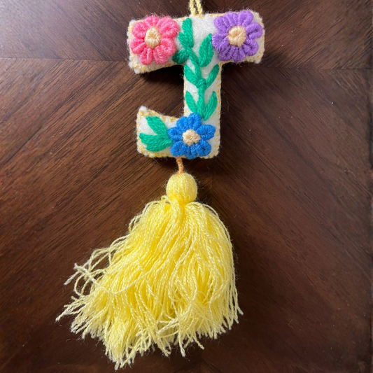 Handmade embroidered letter F ornament with floral patterns and yellow tassel on wooden background