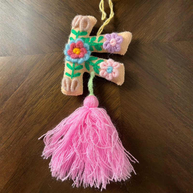 Handmade embroidered letter F ornament with floral patterns on beige fabric and pink tassel on wooden background