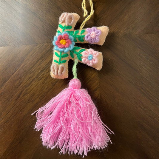 Handmade embroidered letter F ornament with floral patterns on beige fabric and pink tassel on wooden background