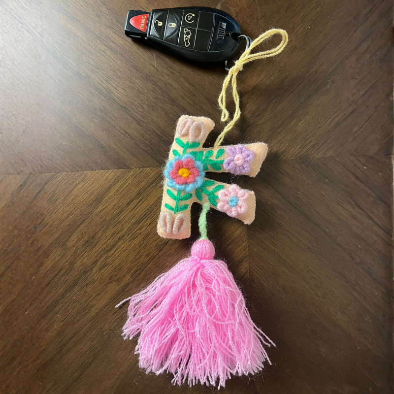 Handmade embroidered letter F ornament with floral patterns on beige fabric and pink tassel on wooden background