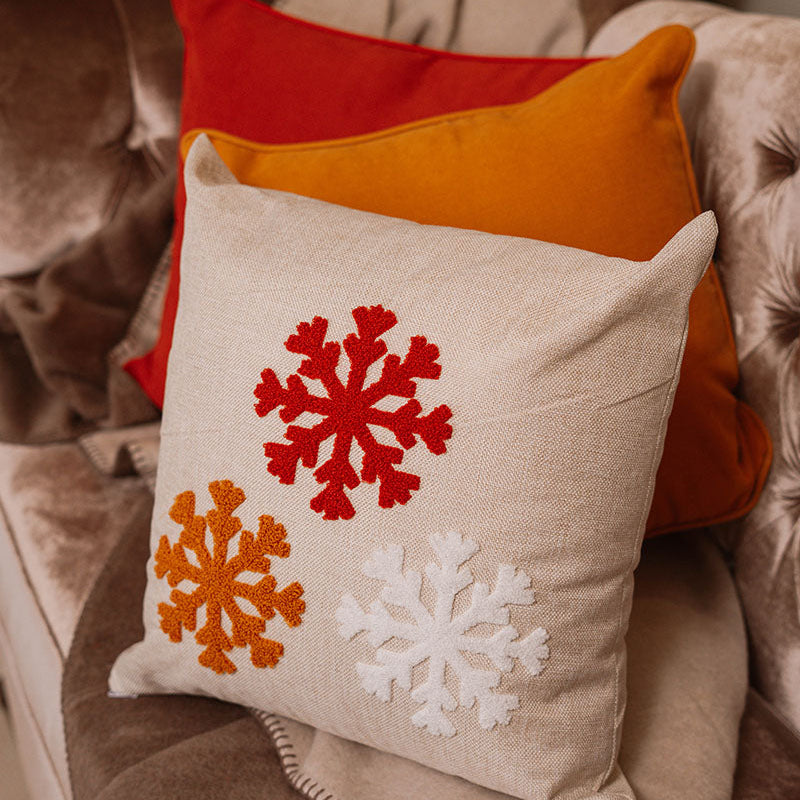 Beige cushion with embroidered snowflakes in white, red, and orange on a blue sofa – Autumn Bloom Collection