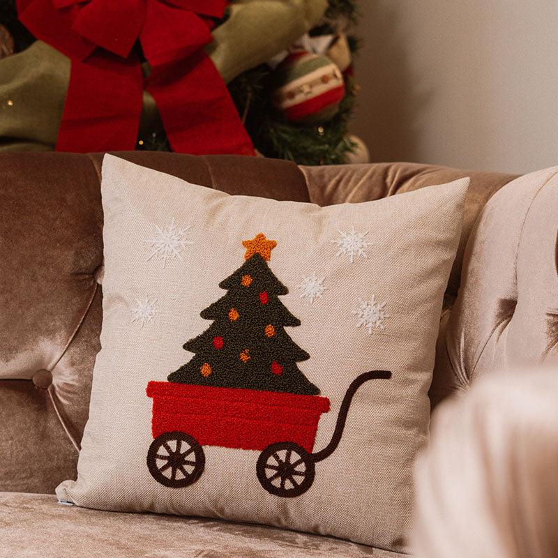 Beige cushion with embroidered red wagon carrying a Christmas tree topped with a gold star on a blue sofa – Autumn Bloom Collection