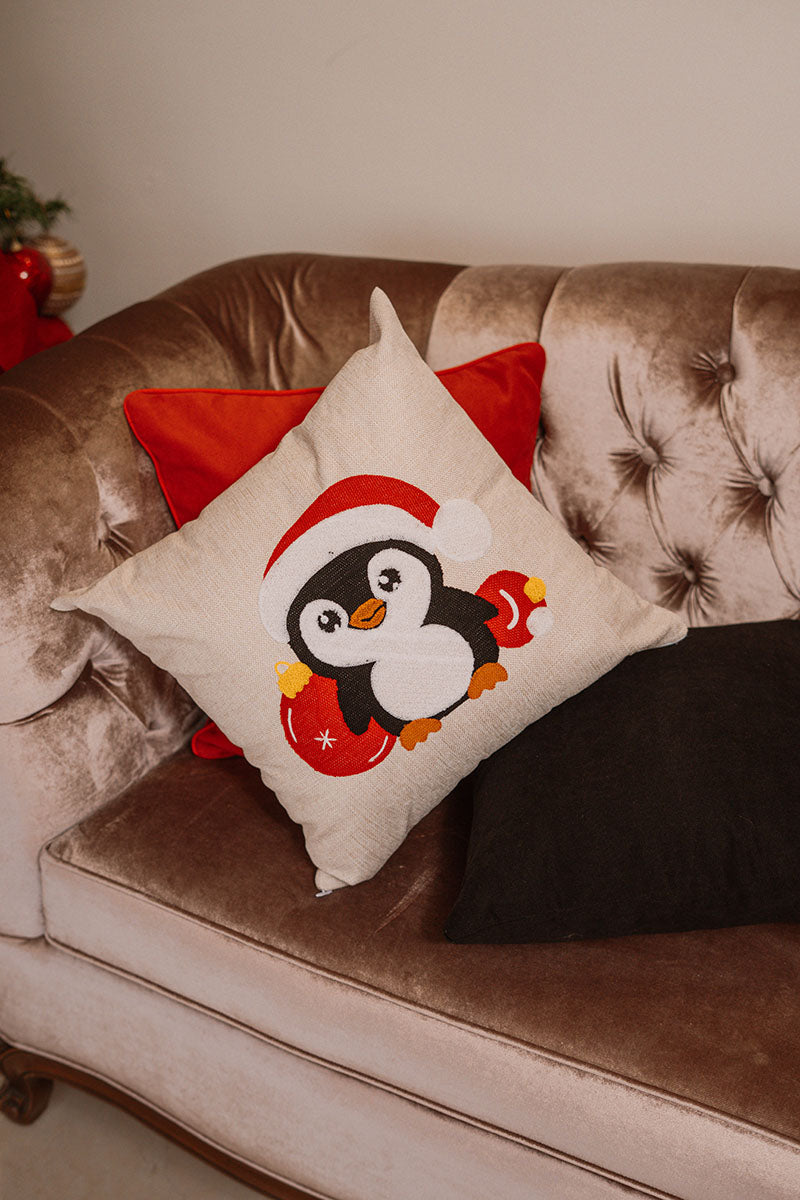 Beige cushion with embroidered Christmas penguin wearing a Santa hat on a blue sofa – Autumn Bloom Collection