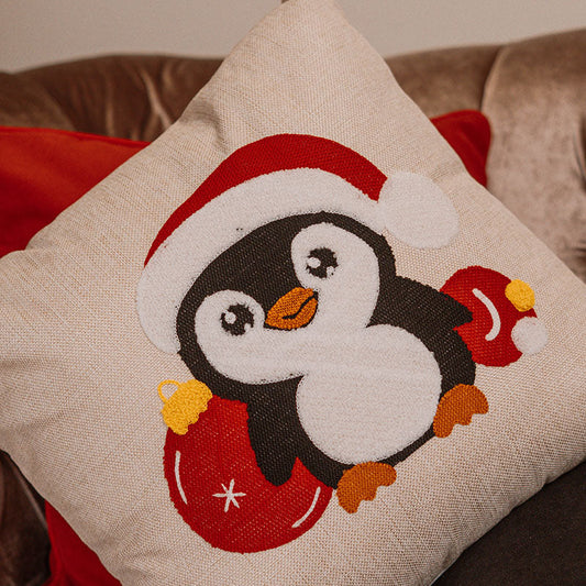 Beige cushion with embroidered Christmas penguin wearing a Santa hat on a blue sofa – Autumn Bloom Collection