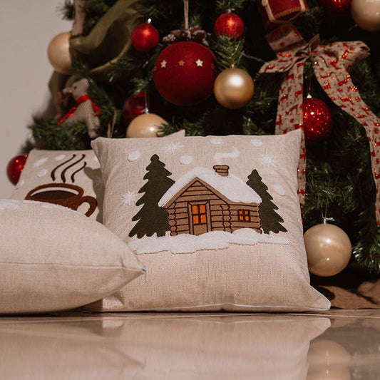 Beige cushion with embroidered winter cabin, snow, and pine trees on a blue sofa – Autumn Bloom Collection