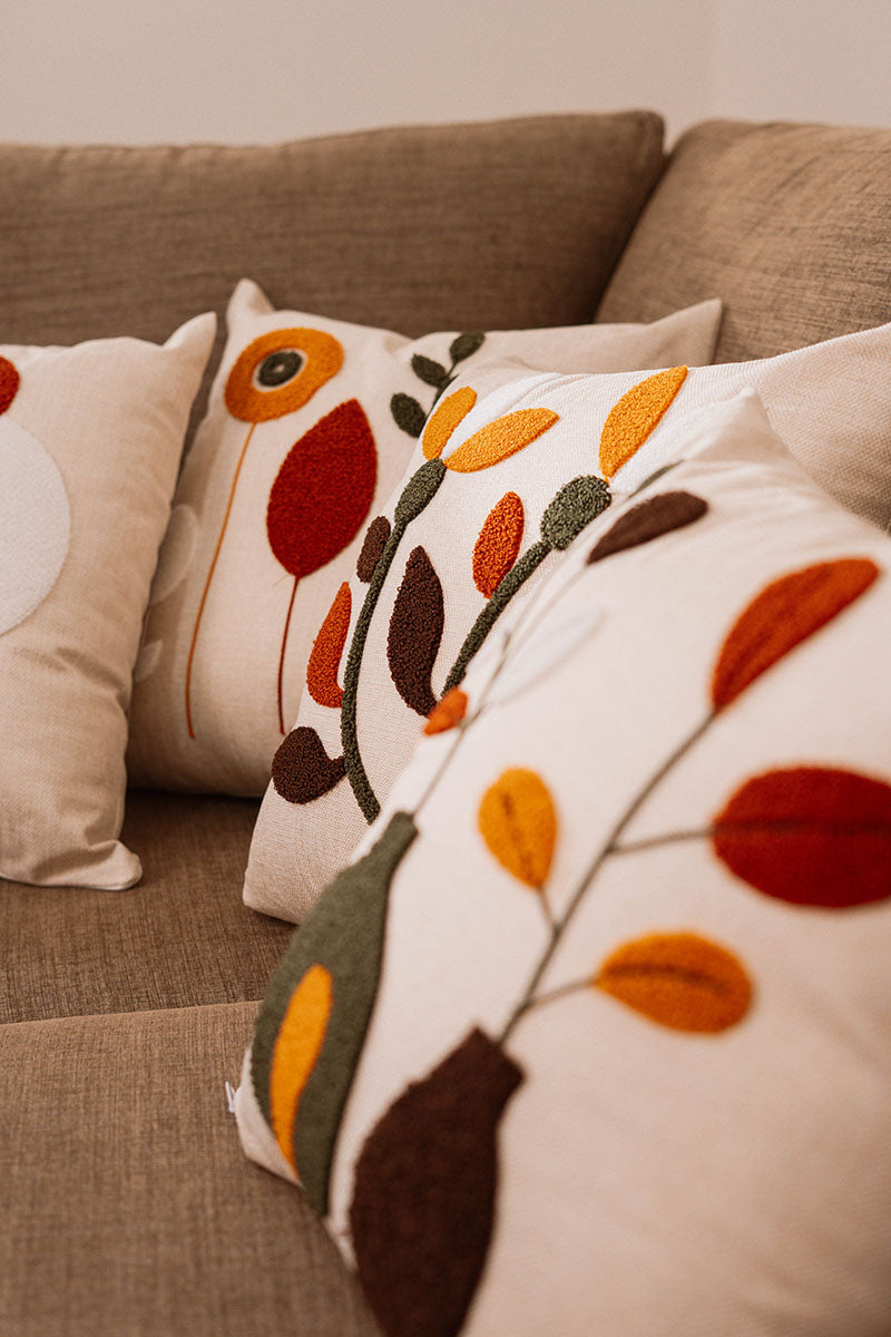 Beige cushion with geometric floral embroidery in rust, mustard, olive, and white on a blue sofa – Autumn Bloom Collection