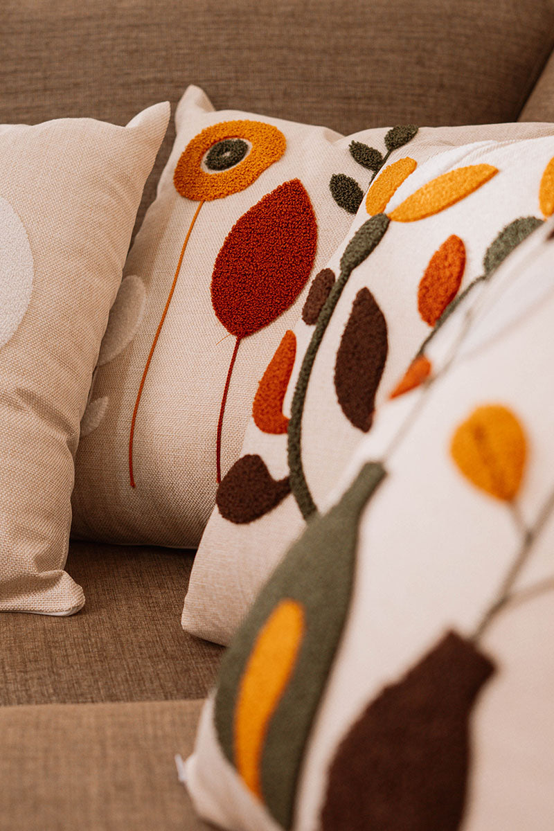 Beige cushion with geometric floral embroidery in rust, mustard, olive, and white on a blue sofa – Autumn Bloom Collection