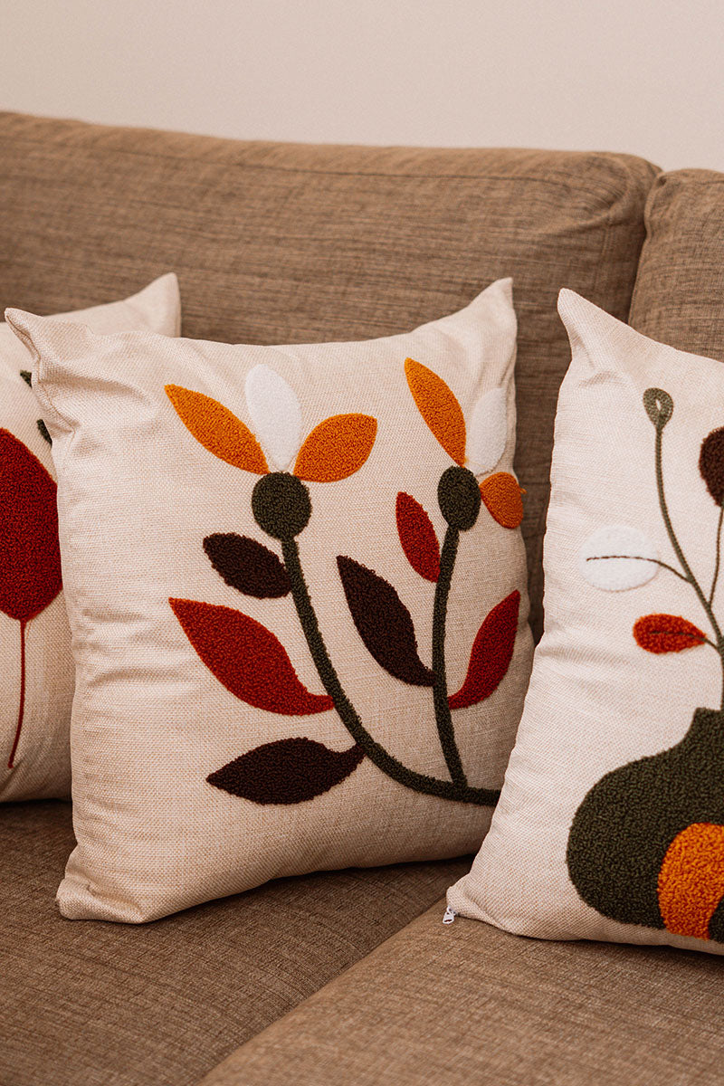 Minimal embroidered floral cushion cover with mustard, rust, and olive leaves on beige fabric, placed on blue sofa