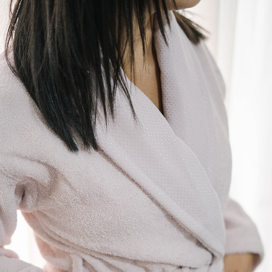 Unisex cotton terry bathrobe with shawl collar and waist belt, worn by woman near window in natural light