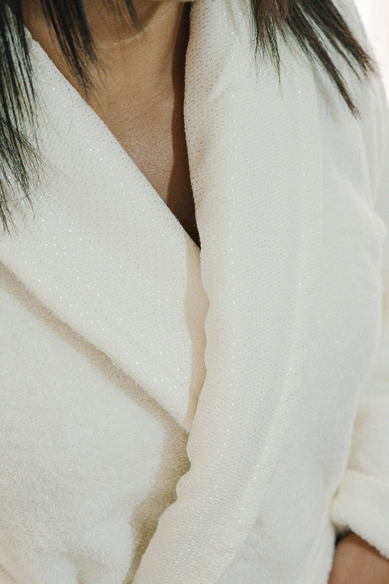 Unisex cotton terry bathrobe with shawl collar and waist belt, worn by woman near window in natural light
