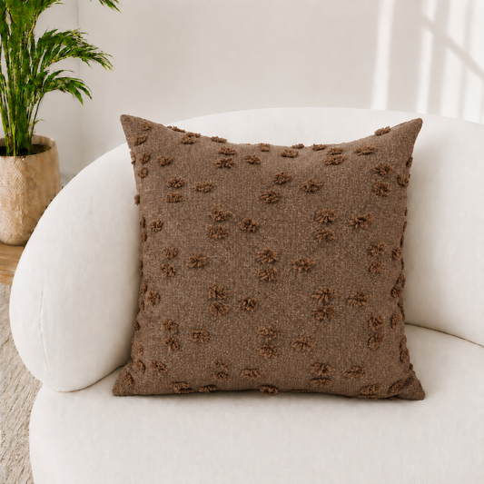 A square brown textured throw pillow with raised pom-pom details, resting on a white curved armchair in a bright, modern room with a potted plant in the background.