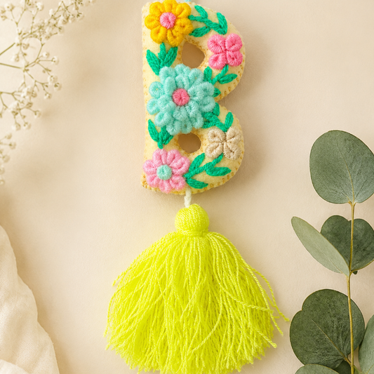 A handmade felt letter 'B' ornament decorated with colorful flowers and a bright yellow tassel, displayed on a beige background with eucalyptus leaves.