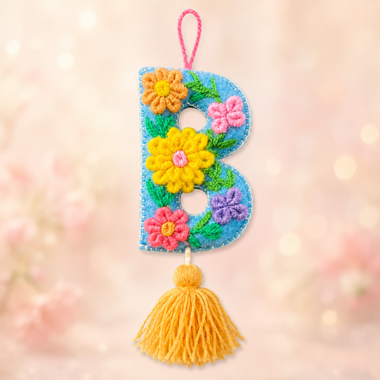 Decorative letter 'B' with flowers and a yellow tassel on a wooden surface