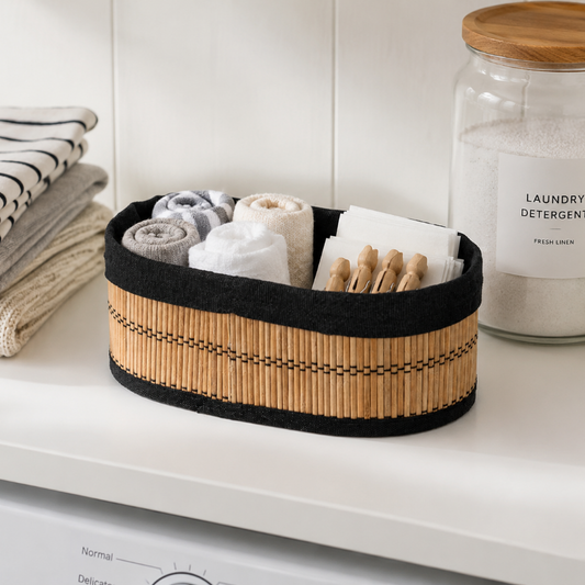 A woven storage basket with a black fabric trim and a natural bamboo slat body, holding rolled towels and fabric swatches, styled on a white shelf next to laundry and detergent jars.