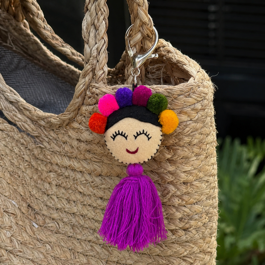 Handmade smiling Frida-inspired doll charm with colorful pom-poms and purple tassel on teal bag