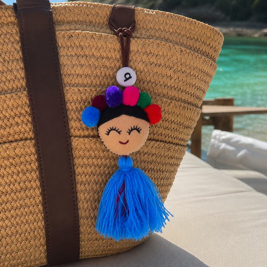 Handmade smiling Frida-inspired doll charm with colorful pom-poms and blue tassel on teal bag