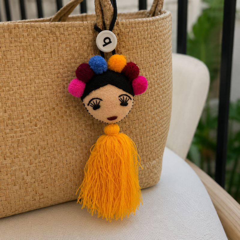 Handmade Frida-inspired doll charm with colorful pom-poms and yellow tassel on teal bag