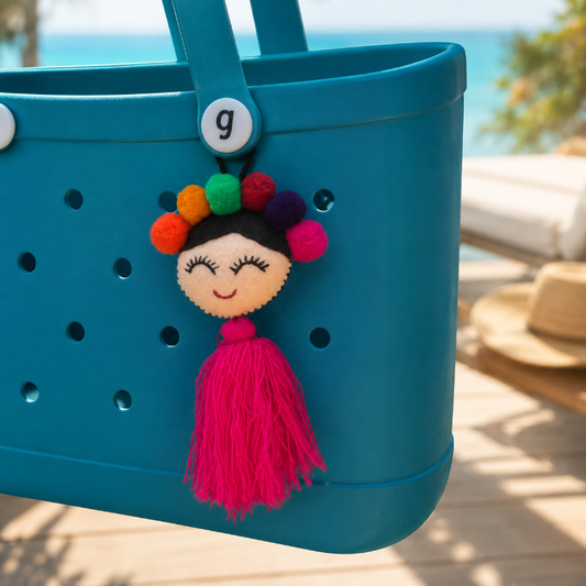 Handmade smiling Frida-inspired doll charm with colorful pom-poms and pink tassel on teal bag