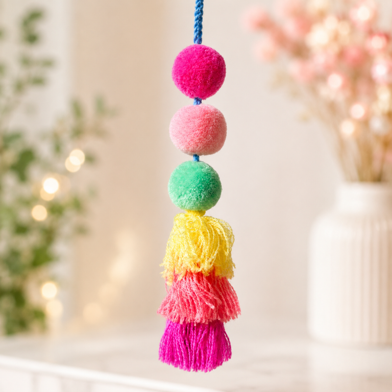 A colorful hanging ornament with four fuzzy pom-poms in pink, green, yellow, and magenta, attached to a blue cord, shown against a softly blurred indoor background.