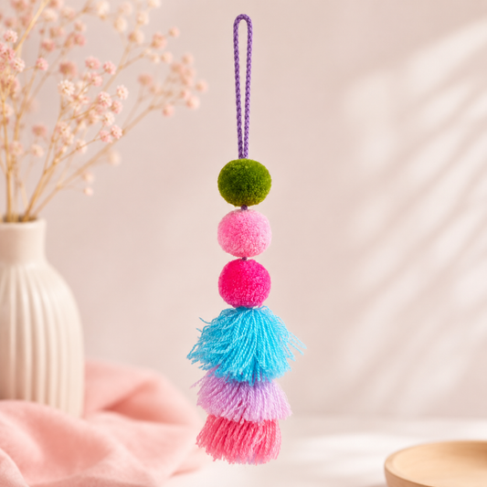 A hanging decorative ornament with a purple loop at the top, three stacked pom-poms in green, pink, and magenta, and a tassel section with blue, lavender, and pink fringe. The ornament is photographed in a softly styled indoor setting with a vase of dried flowers and pink fabric in the background.