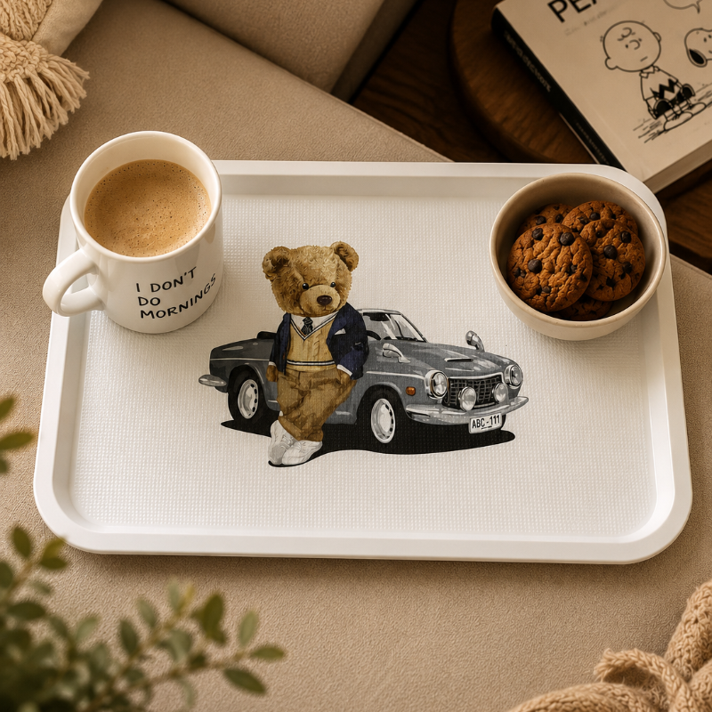 A white rectangular tray styled on a beige sofa, printed with an illustration of a teddy bear in a suit sitting in a gray car. The tray is accompanied by a white mug reading 'I DON'T DO MORNING' with coffee, a small bowl of chocolate chip cookies, and a Peanuts book in a cozy indoor setting.