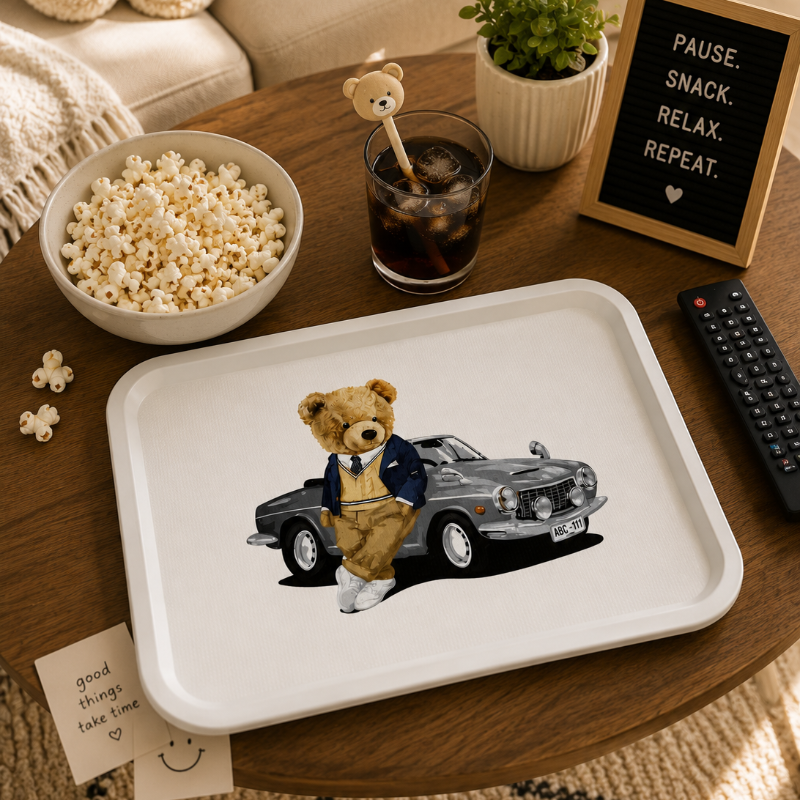 A white rectangular serving tray with rounded corners sits on a wooden table in a cozy living room setting. The tray is printed with an illustration of a teddy bear wearing a navy blue suit and white shoes, standing next to a gray classic car.