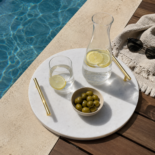 A round white marble serving tray on a wooden deck beside a swimming pool, styled with a clear glass carafe of water with lemon slices, a glass of water with a lemon slice, a small bowl of green olives, gold-toned straws, and a beige fringed throw blanket with sunglasses.