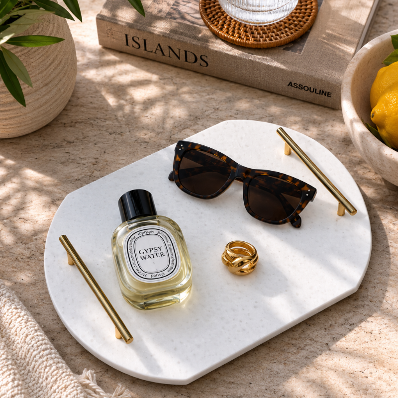 A styled flat-lay featuring a white speckled ceramic tray holding a bottle of Diptyque Cyprus Water perfume, tortoiseshell sunglasses, gold-toned accessories, and a gold ring, set on a textured beige surface with dappled sunlight.