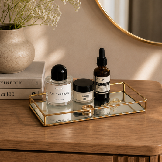 A styled beauty and fragrance setup on a wooden vanity tray, featuring a Rinspo Sal d'Apfousse perfume bottle, a L'Envers skincare jar, and an Aesop dropper bottle, alongside a vase of baby's breath and books.