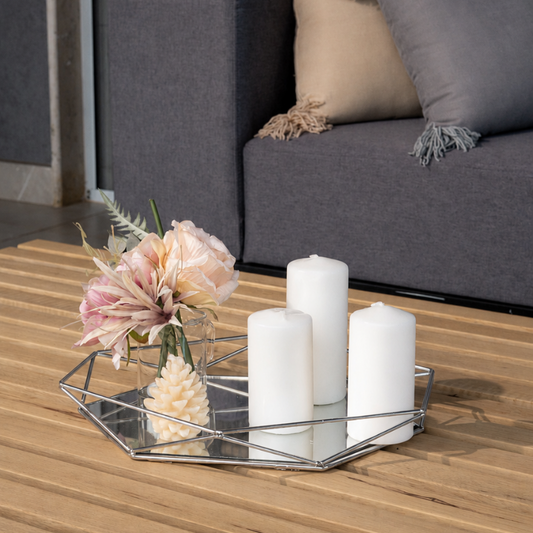A modern geometric mirrored tray on a wooden deck table, styled with three white pillar candles and a small vase of pink flowers. In the background, a contemporary gray sofa with decorative pillows is visible.