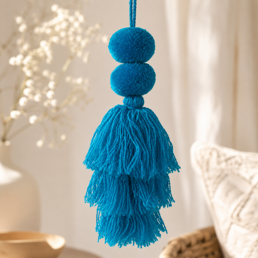 Turquoise blue pom-pom tassel hanging decoration with two pom-poms and layered turquoise yarn tassels hanging on a wooden chair.