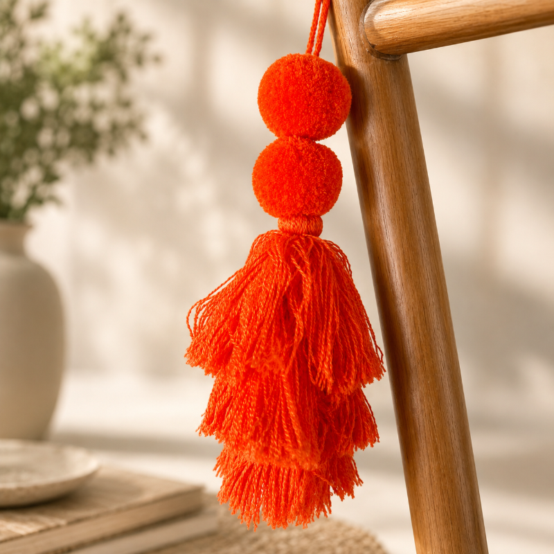 Neon orange pom-pom tassel hanging decoration with two pom-poms and layered neon orange yarn tassels hanging on a wooden chair.