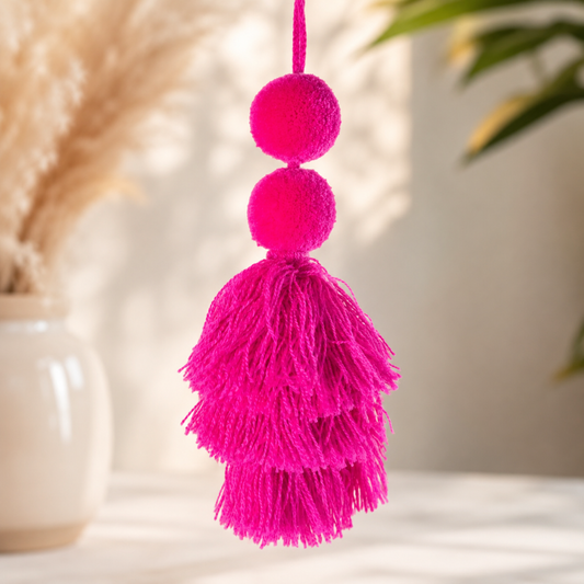 Hot pink pom-pom tassel hanging decoration with two pom-poms and layered pink yarn tassels hanging on a wooden chair.